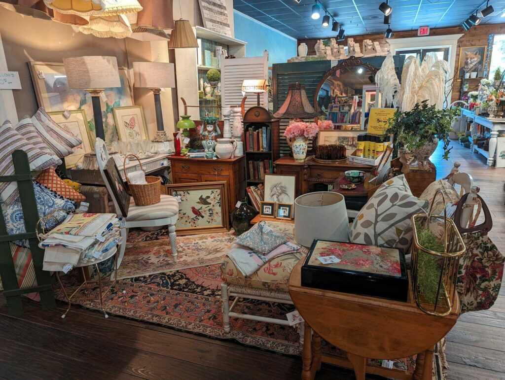 Cozy antique shop display with vintage lamps, furniture, pillows, framed art, and decorative items.