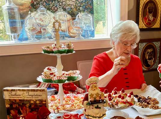 Elderly woman in red prepares desserts on a decorated table with tiered trays and Christmas-themed treats.